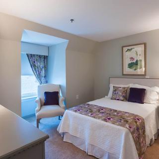 A cozy bedroom with a neatly made bed, an upholstered chair by the window, and wall artwork.