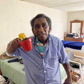 An elderly woman holding a red pot with a yellow flower inside, standing in her living space.
