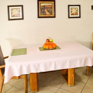 A dining room with a white tablecloth-covered table, adorned with a fruit basket and surrounded by chairs. The walls are decorated with framed artwork.
