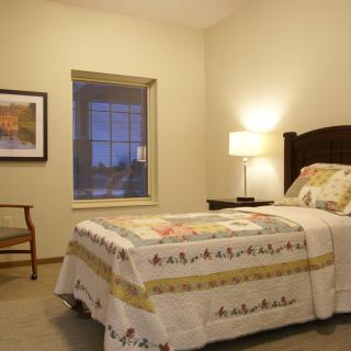 A cozy bedroom with a single bed, a quilted blanket, and a lamp by the window.
