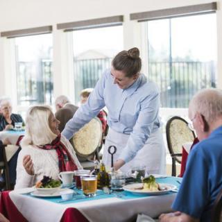 A caregiver assists elderly individuals during a meal at a dining facility.
