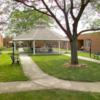 A peaceful outdoor area featuring a gazebo, surrounded by green lawns and pathways, with benches and a brick building in the background.