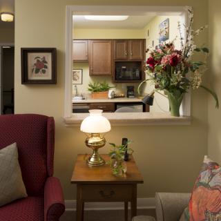 A cozy living room with a view into the kitchen, featuring comfortable furniture, a lamp, and a floral arrangement.