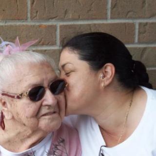 A younger woman affectionately kisses an elderly woman on the cheek in front of a brick wall.