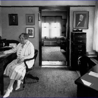 A vintage black and white photo of a woman sitting at a desk in an office, surrounded by portraits on the walls.