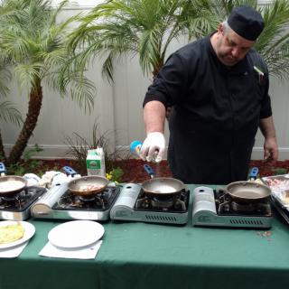 A chef preparing food outdoors using portable stoves surrounded by palm trees.