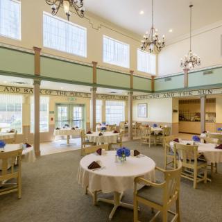 A spacious and elegantly designed dining room with round tables, wooden chairs, and large windows allowing natural light to fill the space.