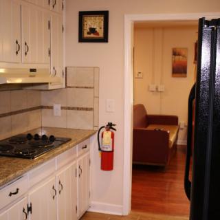 A view of a kitchen with cream cabinets and a black refrigerator, leading to a living room with a brown couch.