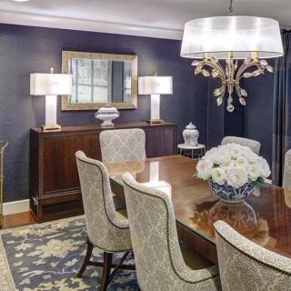 An elegantly designed dining room featuring a large wooden table, upholstered chairs, and a chandelier. The walls are adorned with a mirror and decorative lighting.
