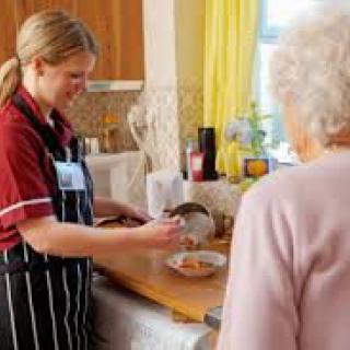 A caregiver assists an elderly person by preparing a meal in the kitchen.