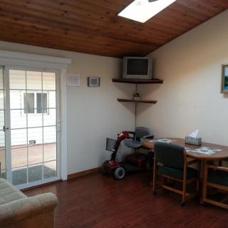 A cozy interior room featuring a wooden ceiling, a small television on a wall-mounted shelf, a mobility scooter parked next to a desk with chairs, and a sliding glass door leading outside.