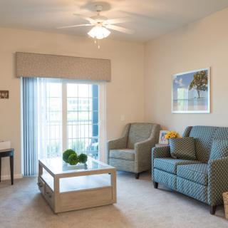 A cozy living room with a sofa and armchair, featuring a central coffee table and a large window with curtains.