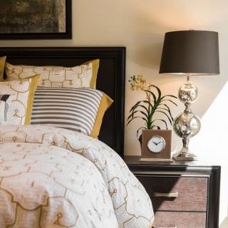 A cozy bedroom scene featuring a bed with patterned pillows and comforter, a nightstand with a clock, lamp, and decorative plant.