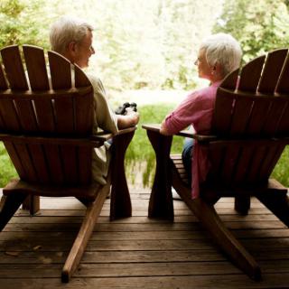 An elderly couple sitting in wooden chairs on a deck, enjoying a peaceful outdoor setting.