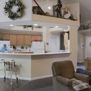 A cozy living area with a view of the kitchen, featuring a wreath decoration on the wall, comfortable seating, and mobility aid equipment.