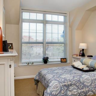 A cozy bedroom with a large window, featuring a bed with patterned bedding and a small lamp on the nightstand.