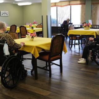 Elderly individuals seated in a dining area of a care facility, with vibrant yellow tablecloths and floral centerpieces.