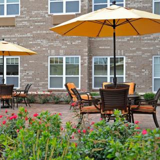 A serene outdoor patio area with tables, chairs, and umbrellas in front of a brick building, surrounded by flowers.