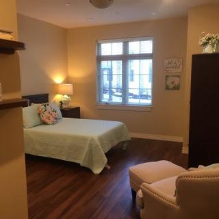 A cozy bedroom with soft lighting, featuring a bed, chair, and a window for natural light.