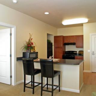 A modern apartment kitchen with an open layout, featuring bar stools and wooden cabinetry.