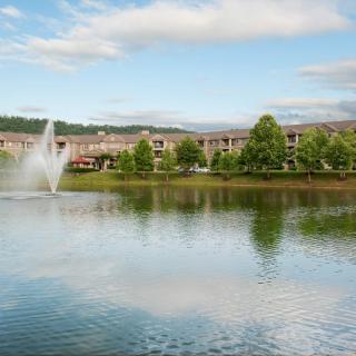 A scenic view of a lake with a fountain, surrounded by trees and buildings under a partly cloudy sky.