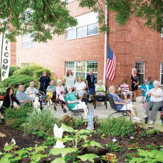 A community event with a group of seniors gathered in a garden area outside a building, featuring an American flag.