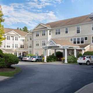 A residential building with a parking lot and driveway, surrounded by trees and under a partly cloudy sky.