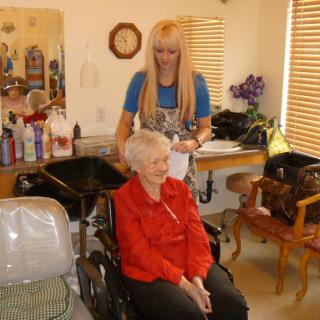 An elderly woman in a wheelchair smiles while getting her hair styled at a salon.