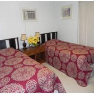 A cozy bedroom featuring two twin beds with patterned bedspreads, a nightstand with lamps and flowers, and framed artwork on the walls.