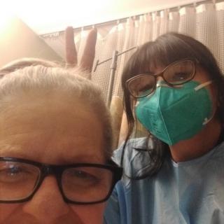 A person taking a selfie with another individual in a medical setting, both wearing glasses and one wearing a mask.