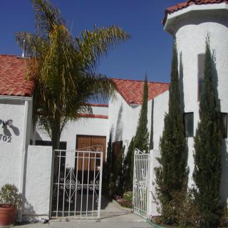 A Mediterranean-style house with white walls and a red-tiled roof, featuring a gated entrance and palm trees.