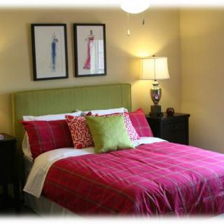 A cozy bedroom with a green headboard, red bedding, and colorful pillows, featuring two framed artworks above the bed and a lamp on the bedside table.
