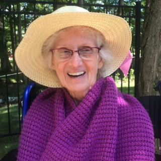 An elderly woman smiling while wearing a large straw hat and a purple shawl, sitting outdoors.