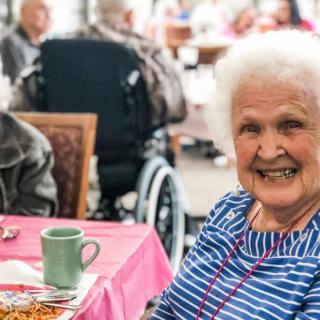 An elderly woman smiling at a dining table in a nursing home.