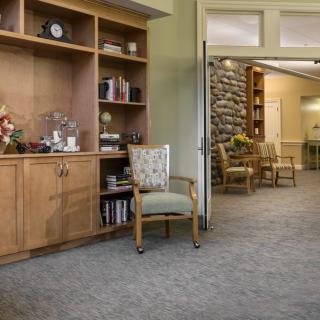 A cozy room with a bookshelf and chairs, featuring warm decor and carpeted flooring.