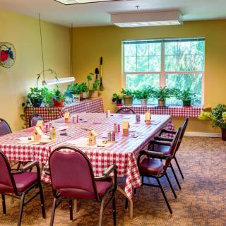 A cozy dining room featuring a large table with a checkered tablecloth, surrounded by chairs and adorned with potted plants near the windows.