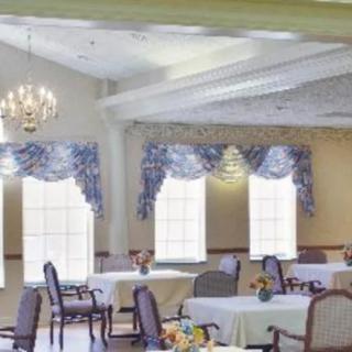 A brightly lit dining room with elegantly set tables and chairs, featuring a chandelier and decorative curtains.