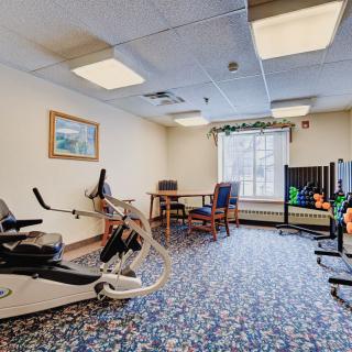 A small exercise room with a stationary bike, weights, and chairs on a carpeted floor.