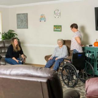 A caregiver and a woman interact with an elderly person in a wheelchair in a living room setting.