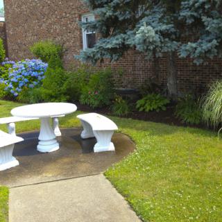 A peaceful garden setting featuring a white stone table with benches surrounded by lush greenery and bushes.