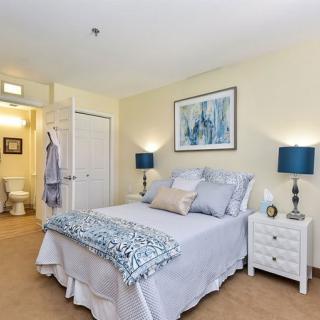 A cozy and elegantly decorated bedroom featuring a neatly made bed with patterned linens, flanked by two white nightstands with blue lamps. A framed artwork hangs above the bed, and an open door leads to a bathroom.