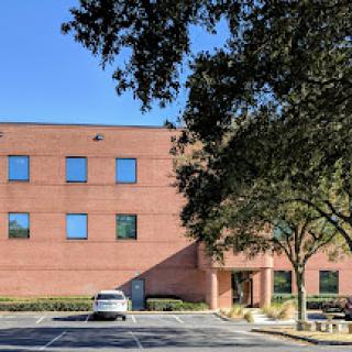 A brick commercial building surrounded by trees and a parking lot on a clear day.