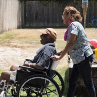 A caregiver assists a person in a wheelchair outdoors on a sunny day.
