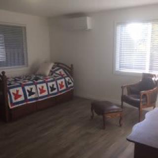 A minimalist bedroom with a daybed featuring a colorful quilt, a wooden chair, and natural light streaming through two windows.