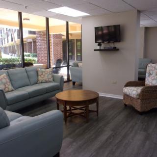 A modern living room with sofas, a coffee table, and a wall-mounted TV, featuring large windows that let in natural light.