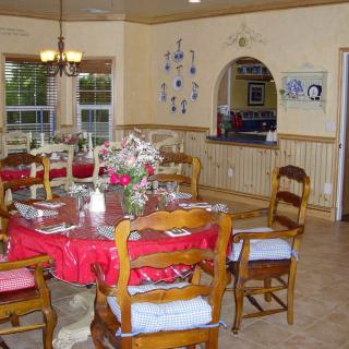 A cozy dining room featuring a round table set with a red tablecloth and floral centerpiece, surrounded by wooden chairs under a chandelier.
