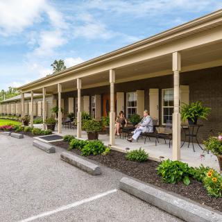 A view of a motel with people sitting on the porch, surrounded by a parking lot and greenery.