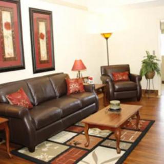 A cozy living room featuring a dark leather sofa and armchair, complemented by red accent pillows and matching wall art. The space is completed with a wooden coffee table, a standing lamp, and an indoor plant.