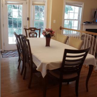 A dining room with a table set with a white tablecloth and flower centerpiece, surrounded by chairs and illuminated by natural light from the windows.