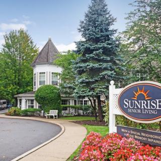 A picturesque building with surrounding trees and flowers, featuring a 'Sunrise Senior Living' sign by the pathway.